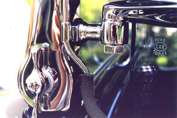 This close-up shows the earlier type safety glass marking. It is a scaled down (smaller) version of windshield markings. The original vacuum hose is cloth reinforced and has a cloth impression on the exterior. Like the terne finish vacuum tubing used elsewhere, the chromed tube on the stanchion has a lengthwise seam from the wrapped construction. The bright reflections tend to hide the distinct seam, but it can be seen just above the hose in this photo. The windshield and stanchion hinges used a small cadmium plated flat washer under the wingnuts.